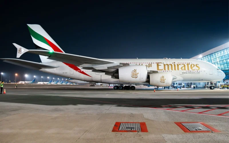 Emirates wide body jet parked at night on an airport tarmac with terminal building in the background