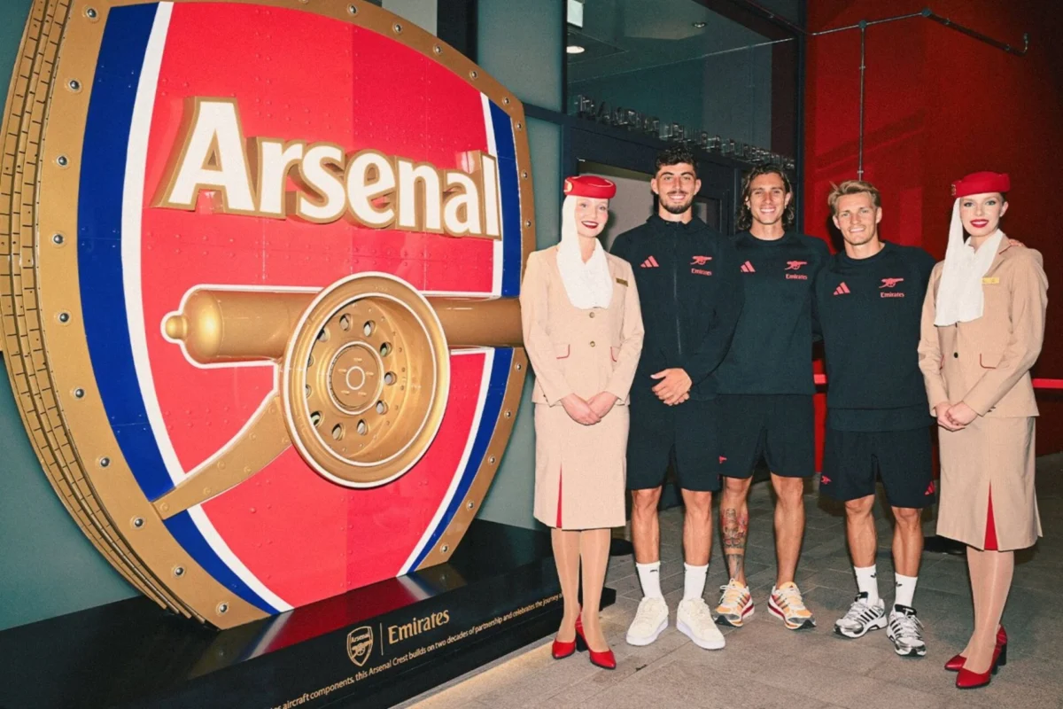 Five peopletwo women in beige outfits and three men in black Emirates gearstand beside a giant red Arsenal vault display