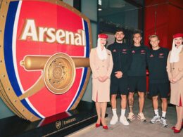 Five peopletwo women in beige outfits and three men in black Emirates gearstand beside a giant red Arsenal vault display