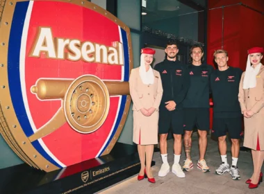 Five people—two women in beige outfits and three men in black Emirates gear—stand beside a giant red Arsenal vault display.