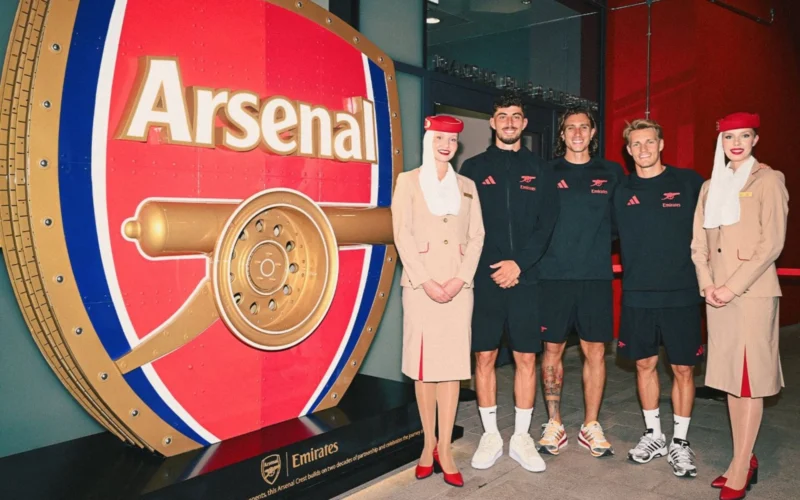 Five peopletwo women in beige outfits and three men in black Emirates gearstand beside a giant red Arsenal vault display