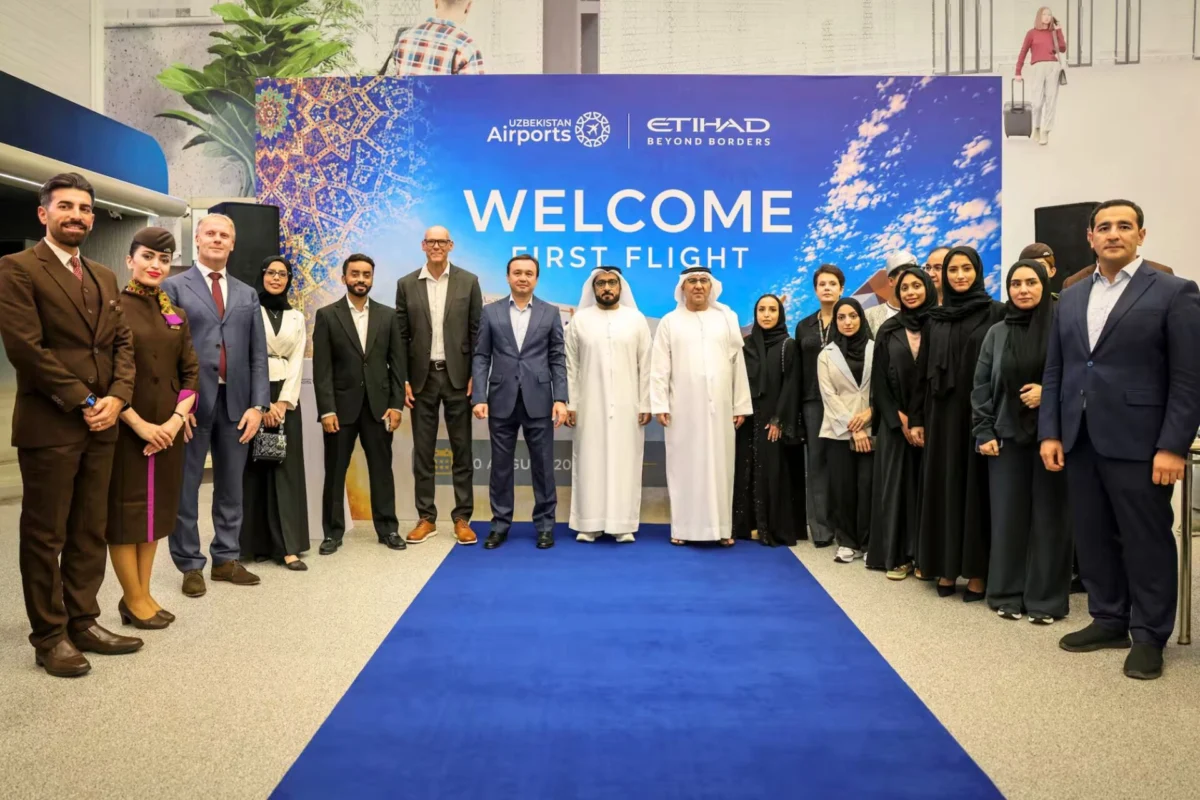 Etihad launches sold out inaugural flight to Tashkent Group of professionals in suits and traditional dress posing on a blue carpet at a Welcome First Flight event in front of a colorful banner