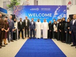 Group of professionals in suits and traditional dress posing on a blue carpet at a Welcome First Flight event in front of a colorful banner