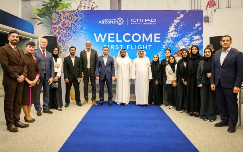 Group of professionals in suits and traditional dress posing on a blue carpet at a Welcome First Flight event in front of a colorful banner