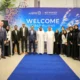 Group of professionals in suits and traditional dress posing on a blue carpet at a Welcome First Flight event in front of a colorful banner