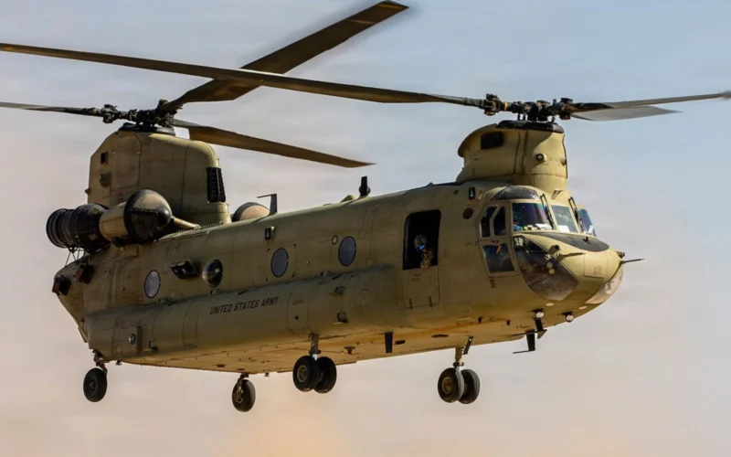 Twin rotor US Army helicopter Chinook in flight side view with open cabin and landing gear visible against a sunset sky