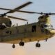 Twin rotor US Army helicopter Chinook in flight side view with open cabin and landing gear visible against a sunset sky