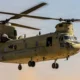 Twin rotor US Army helicopter Chinook in flight side view with open cabin and landing gear visible against a sunset sky