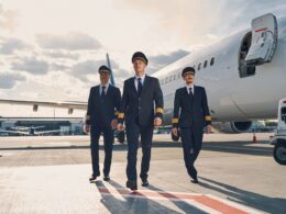 Three pilots in navy uniforms walk toward camera on the airport tarmac beside a large white jet with its door open