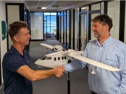 Two men shake hands in a hallway with a white model airplane between them smiling at each other in an office setting