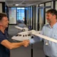 Two men shake hands in a hallway with a white model airplane between them smiling at each other in an office setting