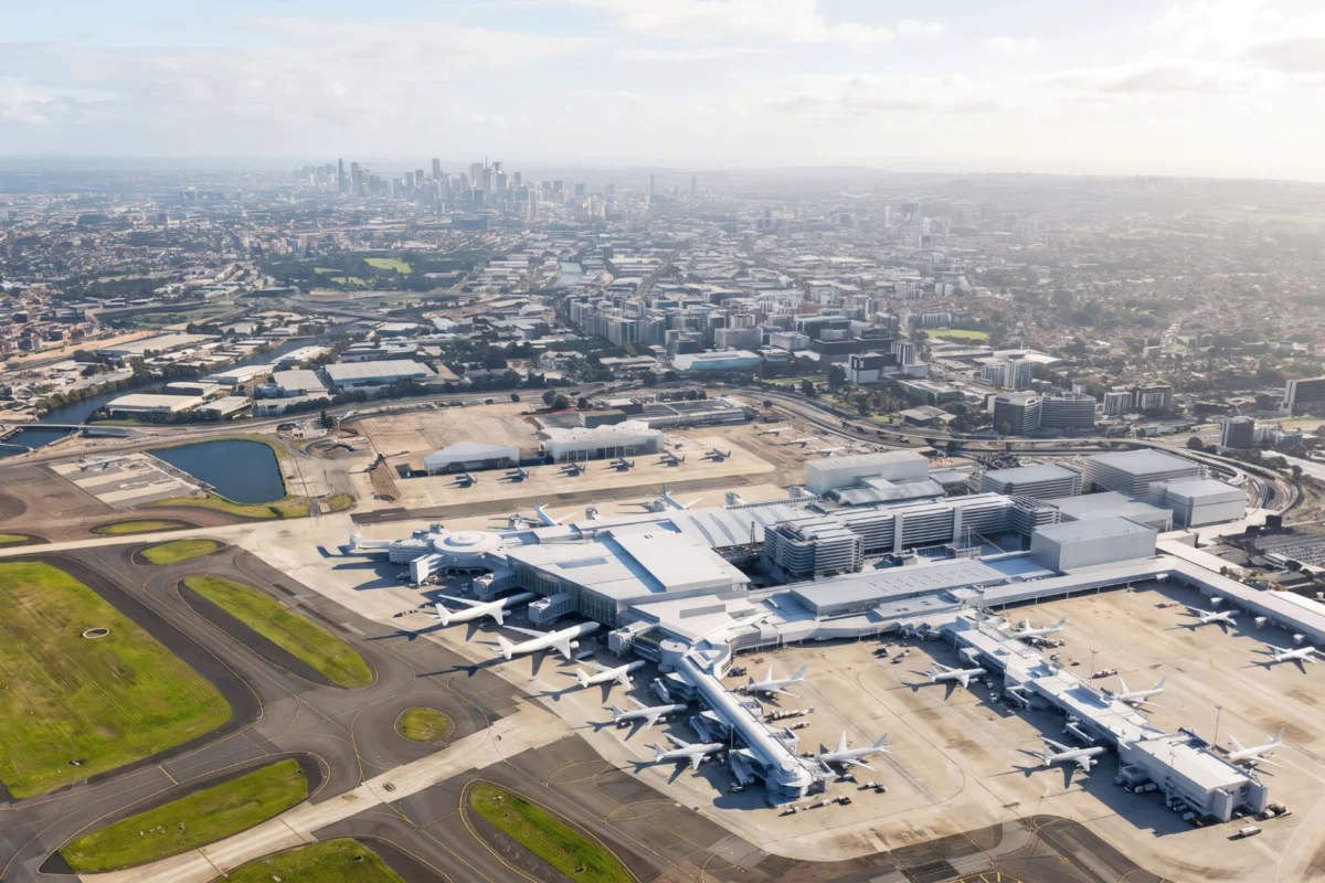 Sydney Airports Master Plan targets integrated terminal Aerial view of a large airport terminal with many airplanes parked at gates and a distant city skyline in the background