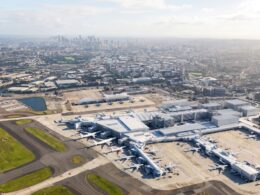 Aerial view of a large airport terminal with many airplanes parked at gates and a distant city skyline in the background