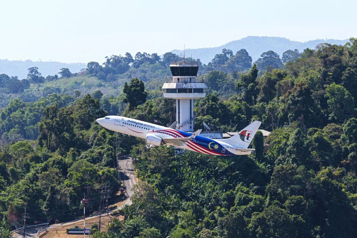 Two Malaysia Airlines jets taking off near a forested airport with a control tower in the background and hills beyond