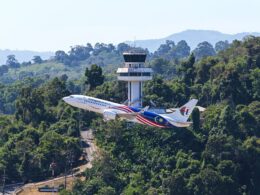 Two Malaysia Airlines jets taking off near a forested airport with a control tower in the background and hills beyond
