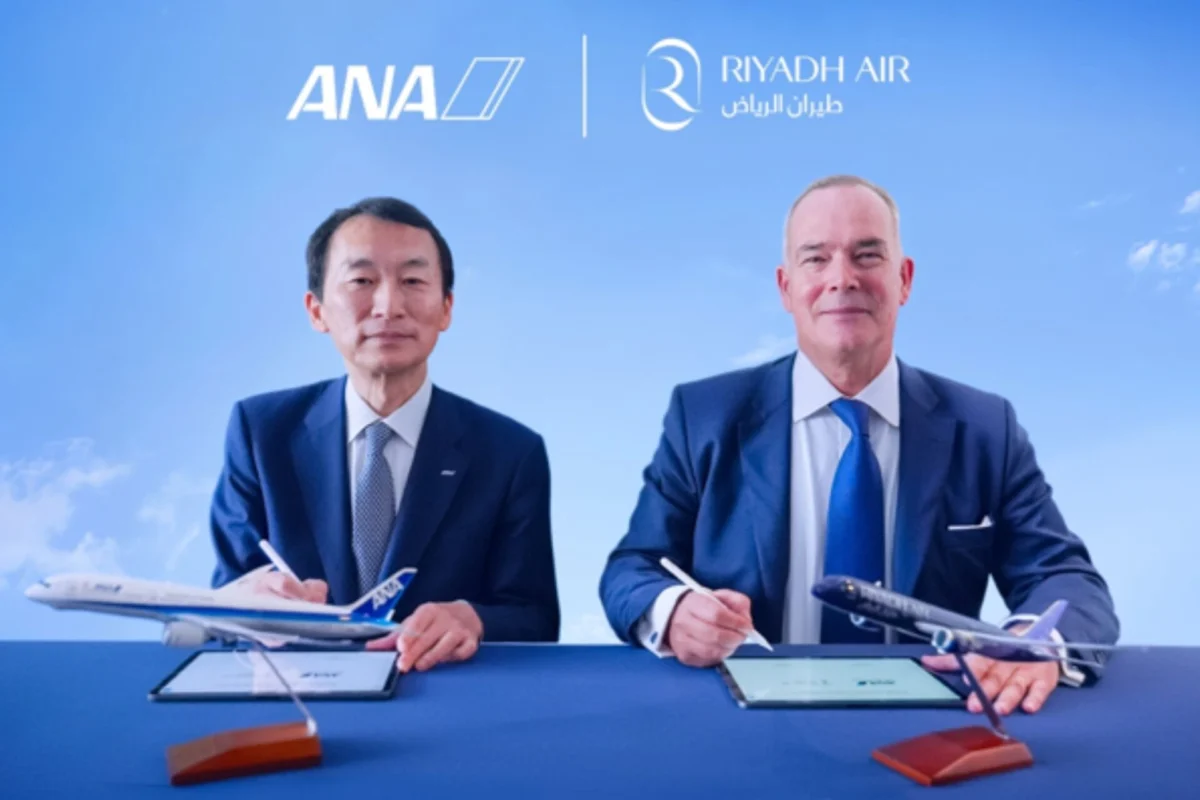 Two men in suits sign documents at a blue table with model planes under ANA and Riyadh Air logos in a clear blue sky backdrop