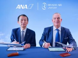 Two men in suits sign documents at a blue table with model planes under ANA and Riyadh Air logos in a clear blue sky backdrop
