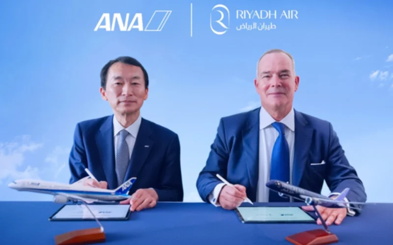 Two men in suits sign documents at a blue table with model planes under ANA and Riyadh Air logos in a clear blue sky backdrop