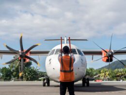Ground crew member in an orange safety vest stands facing a twin engine turboprop aircraft on the tarmac preparing for takeoff