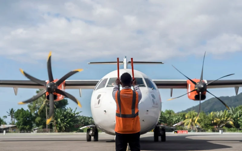 Ground crew member in an orange safety vest stands facing a twin engine turboprop aircraft on the tarmac preparing for takeoff