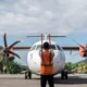 Ground crew member in an orange safety vest stands facing a twin engine turboprop aircraft on the tarmac preparing for takeoff