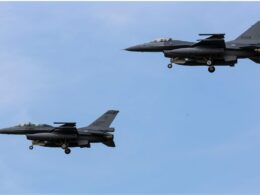 Three gray fighter jets in formation mid flight against a clear blue sky their landing gears down as they pass overhead