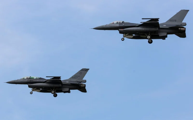 Three gray fighter jets in formation mid flight against a clear blue sky their landing gears down as they pass overhead