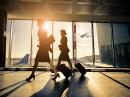 Two silhouette women walking with rolling suitcases through an airport concourse at sunset large glass windows behind them