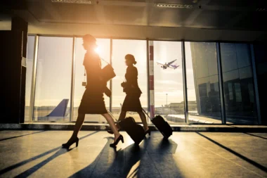 Two silhouette women walking with rolling suitcases through an airport concourse at sunset large glass windows behind them