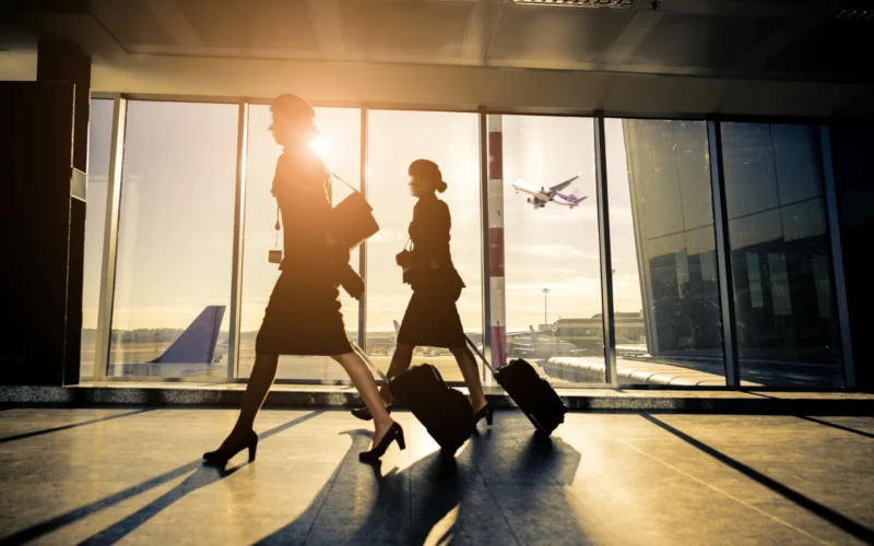 Two silhouette women walking with rolling suitcases through an airport concourse at sunset large glass windows behind them