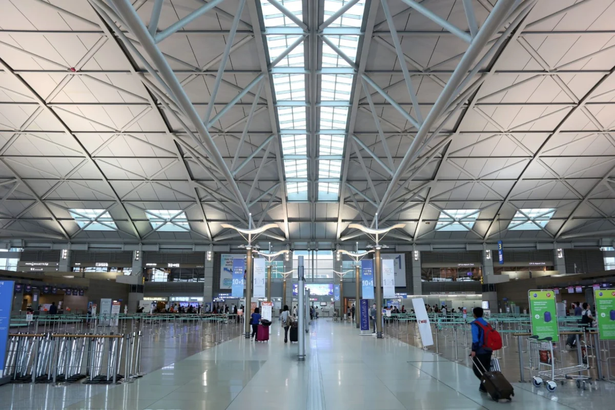 Incheon Airport adds fence after viral queue jumping video Wide view of a spacious airport terminal with a geometric steel beam ceiling skylight panels and travelers with luggage in the foreground