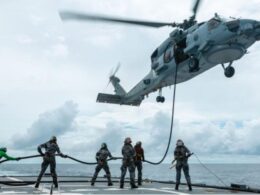 Military helicopter hovers above a ship deck lowering a rescue line to sailors preparing to hoist