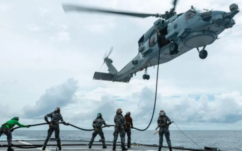 Military helicopter hovers above a ship deck lowering a rescue line to sailors preparing to hoist