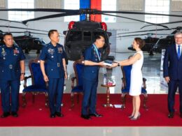 Officials in military uniforms and a woman in a white dress exchange a book in a helicopter hangar with a red carpet backdrop