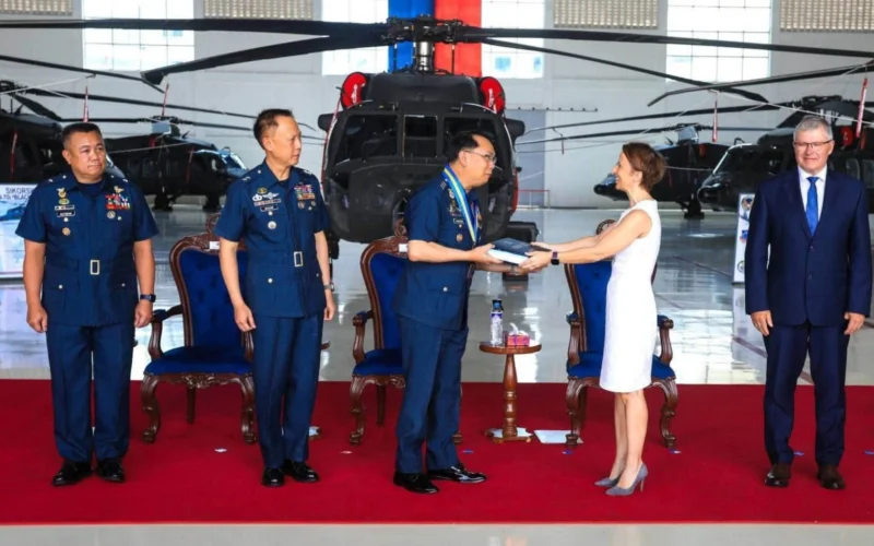 Officials in military uniforms and a woman in a white dress exchange a book in a helicopter hangar with a red carpet backdrop