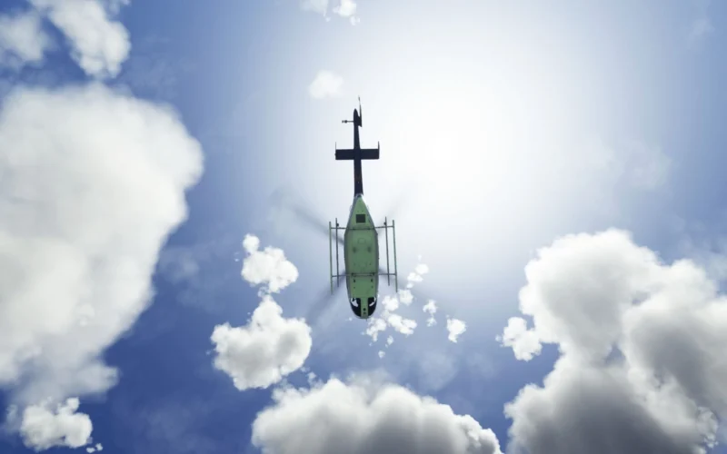 Green helicopter photographed from below against a bright sun and scattered clouds in a blue sky view from beneath the aircraft