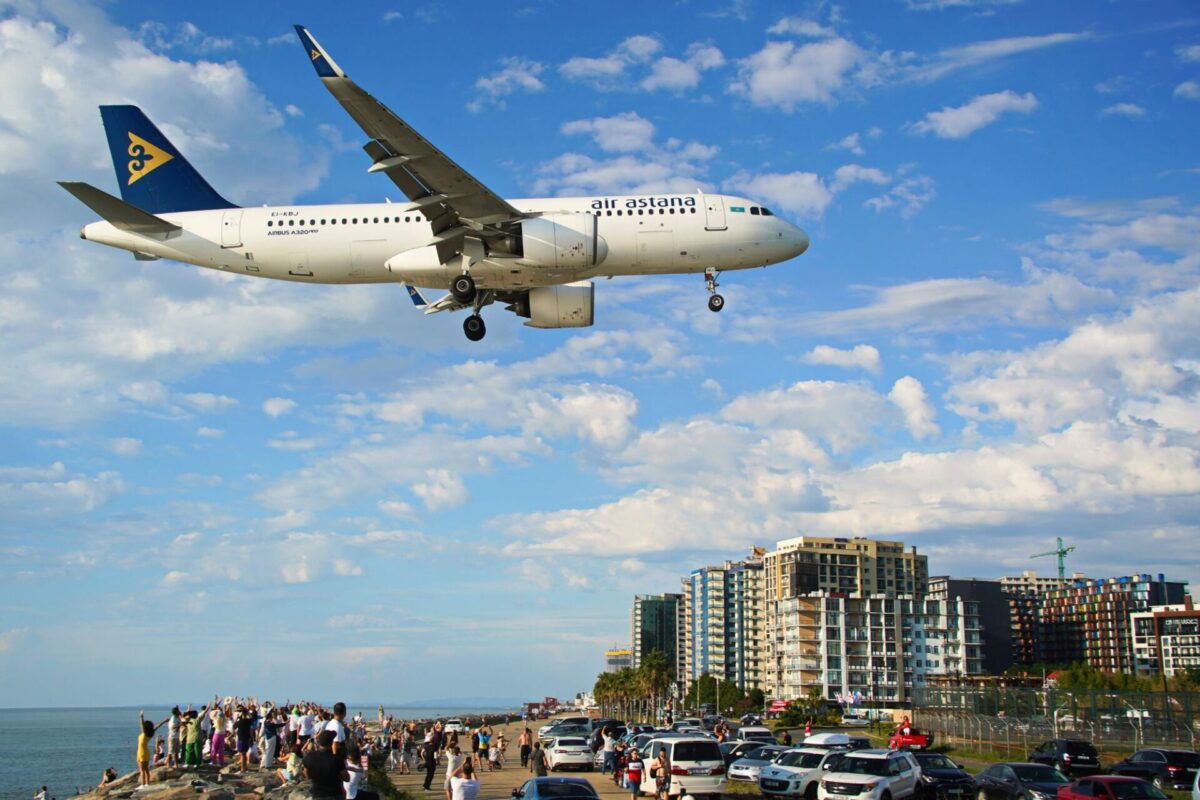 Air Astana Airbus A320neo flying low over a crowded beachfront with people watching and cars parked along the promenade city buildings in the background