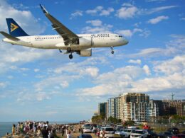 Air Astana Airbus A320neo flying low over a crowded beachfront with people watching and cars parked along the promenade city buildings in the background