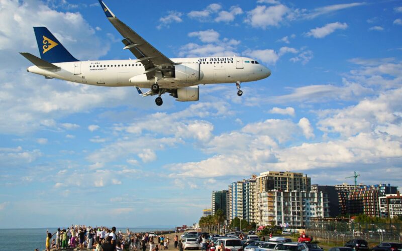 Air Astana posts H1 loss as fuel costs offset revenue gain Air Astana Airbus A320neo flying low over a crowded beachfront with people watching and cars parked along the promenade city buildings in the background