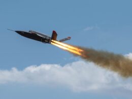 Military jet flying left to right with orange afterburner flames and a smoky trail against a blue sky in flight