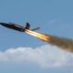 Military jet flying left to right with orange afterburner flames and a smoky trail against a blue sky in flight
