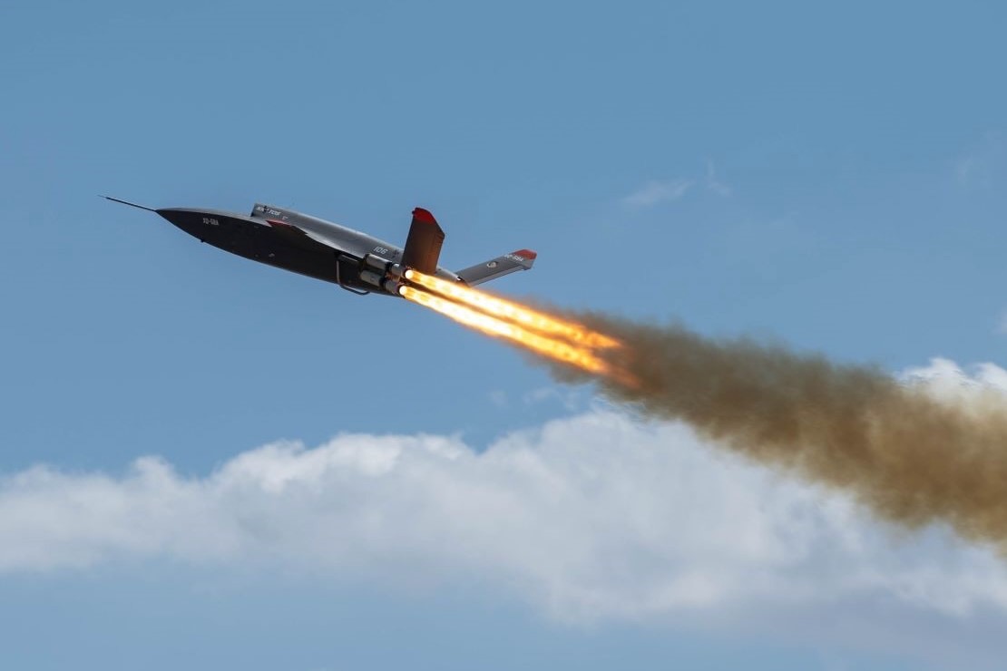 Valk Takeoff Hero Military jet flying left to right with orange afterburner flames and a smoky trail against a blue sky in flight