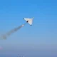 White delta wing aircraft launches a rocket leaving an orange flame and smoke trail over a blue sea and clear sky