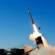 Missile launching from a ground launcher with a bright flame and smoke plume against a blue sky