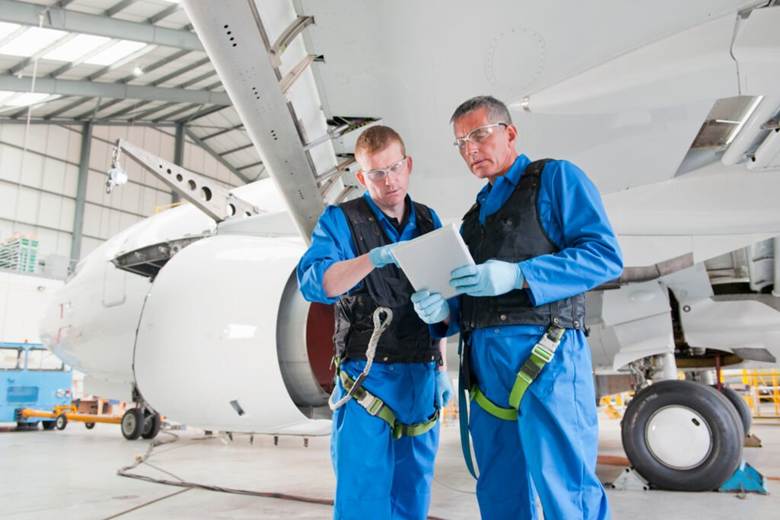 image Two aerospace technicians in blue coveralls and safety harnesses review a tablet beside a large airplane engine in a hangar