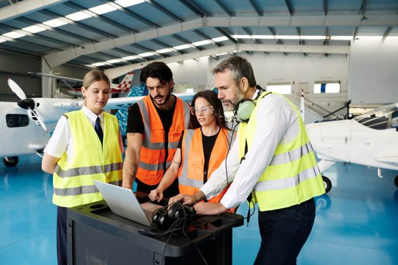 image Four aircraft maintenance workers in safety vests gather around a laptop on a cart inside a hangar inspecting data together