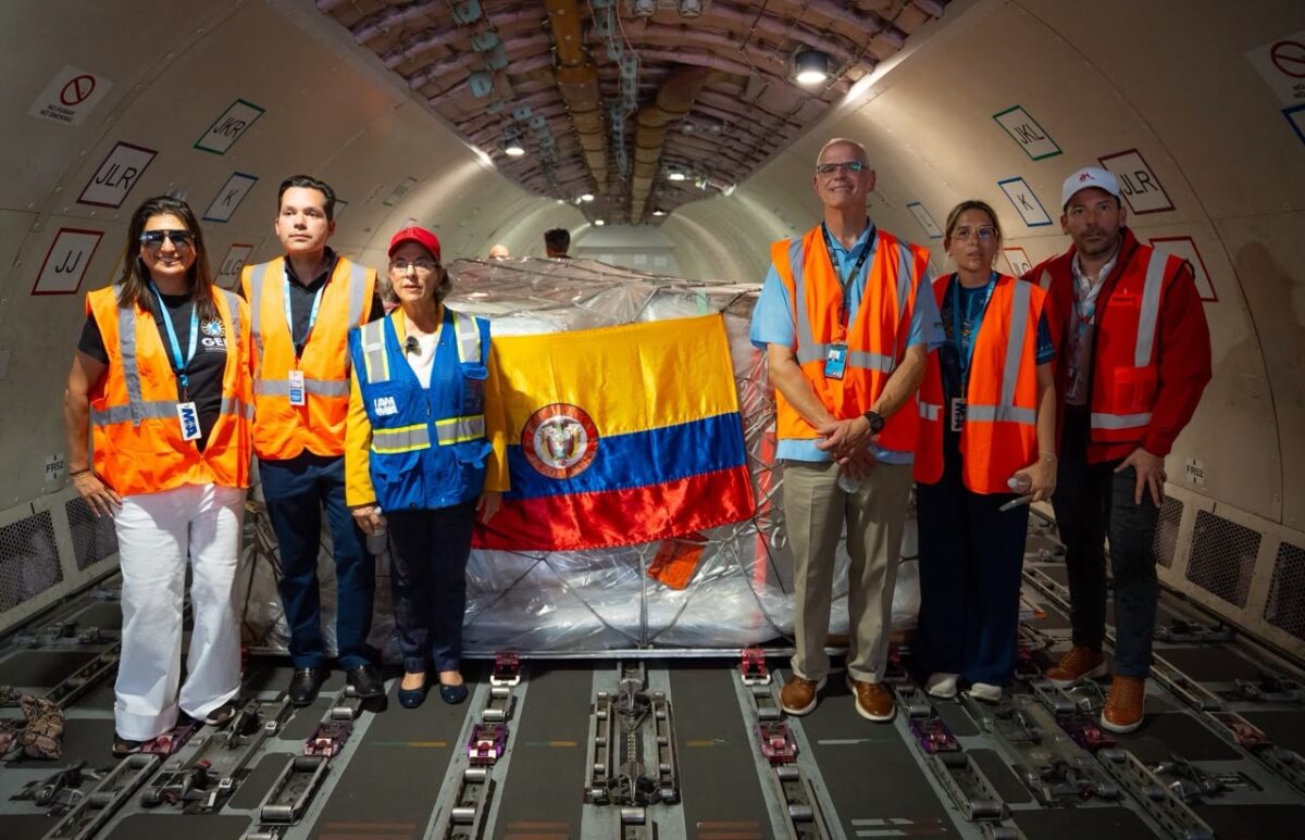 image Seven people in orange safety vests stand in a cargo planes hold a wrapped pallet with the Colombian flag behind them from left to right
