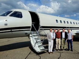 Five men in smart casual attire pose beside a white private jet with its door open and a portable staircase on the tarmac