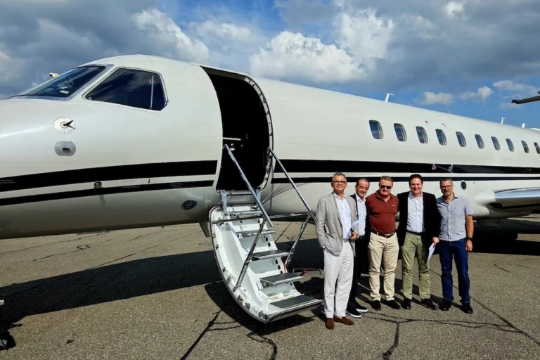 Five men in smart casual attire pose beside a white private jet with its door open and a portable staircase on the tarmac