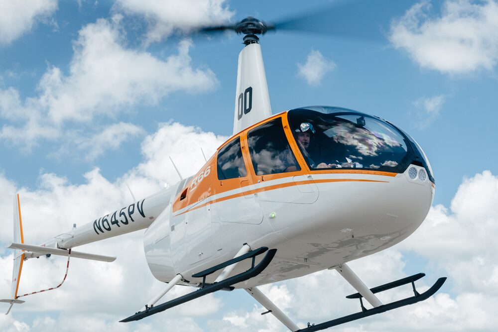r66 White helicopter with an orange stripe flying in a clear blue sky with scattered clouds